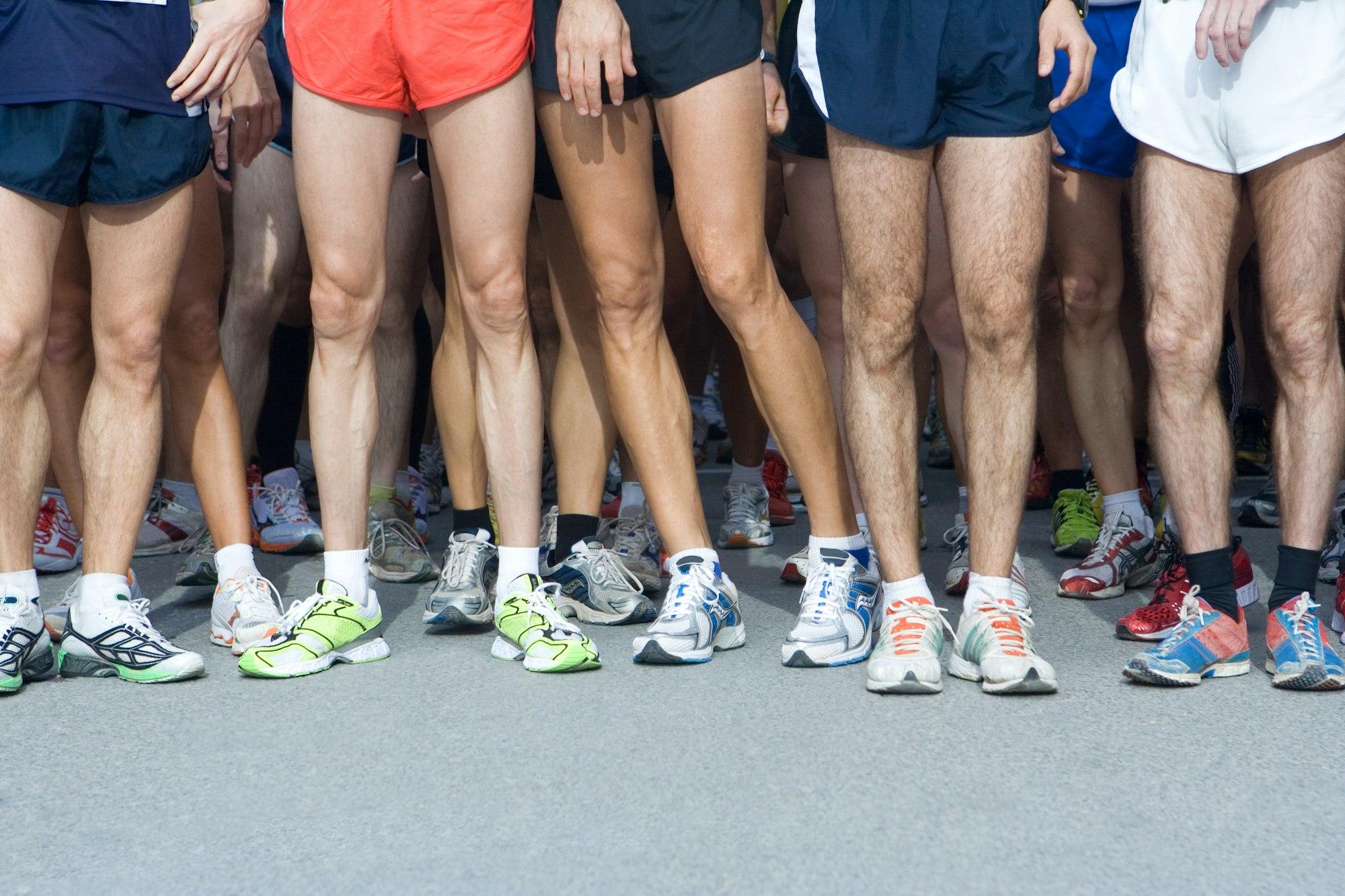 A waist-down photo of a group of runners with bare legs and various running shoes, getting ready to start a race.